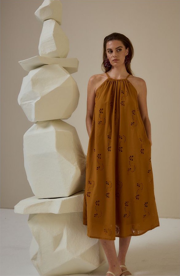 Model in a rust broderie halter midi dress beside a stacked white stone sculpture