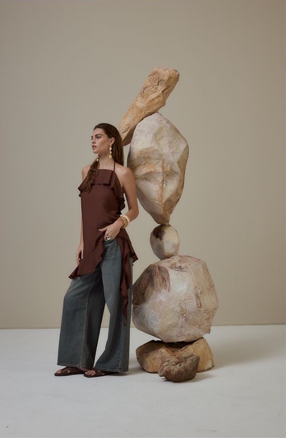 Model in a brown ruffled halter top and wide-leg denim beside a balanced stone sculpture