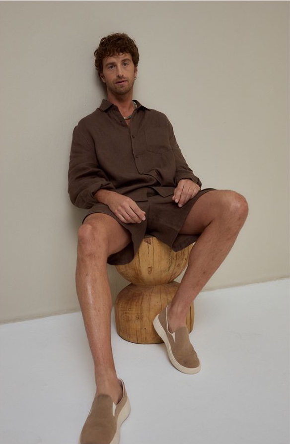 Man in a brown linen shirt and shorts seated on a carved wooden stool